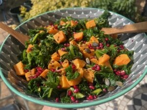 A bowl of salad containing kale, sweet potato cubes, pomegranate seeds, and pumpkin seeds, with wooden utensils, outdoors in natural light.
