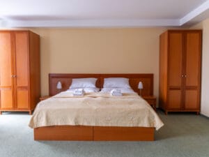 A neatly made double bed with beige bedding, flanked by two wooden wardrobes and bedside tables, in a clean, minimal bedroom.