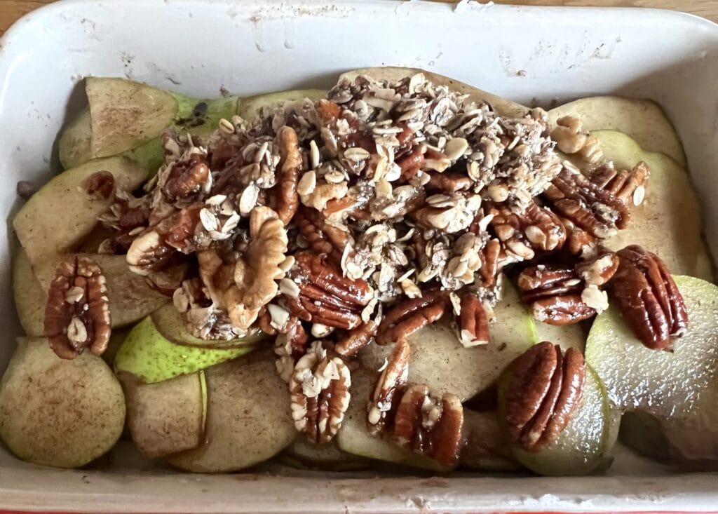 Sliced pears in a baking dish topped with a mixture of rolled oats, pecans, walnuts, and cinnamon, prepared for baking.