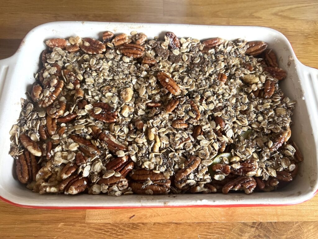 A rectangular baking dish filled with Pear Pecan & Cinnamon Autumn Crumble, topped with a golden oat and pecan topping, sits invitingly on a wooden surface.