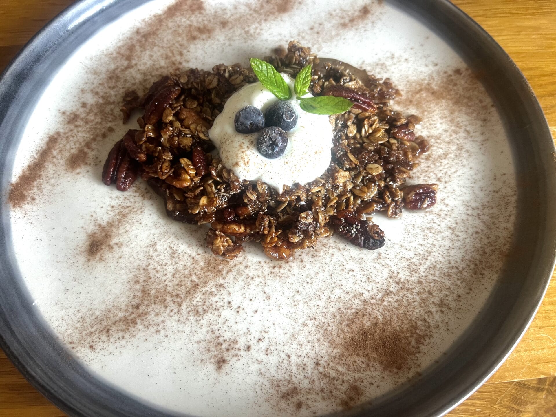 A dessert plate with granola, pecans, and a scoop of ice cream topped with blueberries and mint, garnished with cocoa powder—reminiscent of a Pear Pecan & Cinnamon Autumn Crumble.