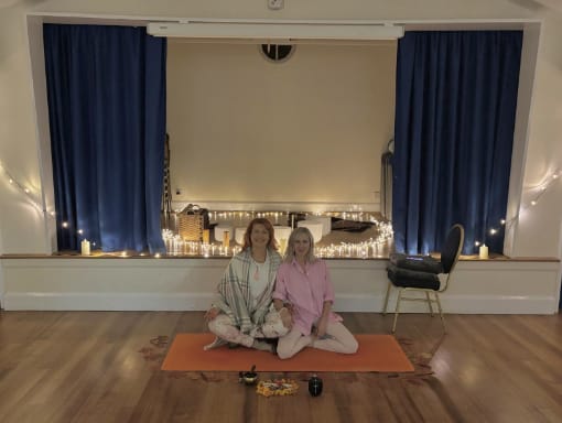 Two women sit cross-legged on an orange yoga mat in a softly lit room with blue curtains, string lights, and candles. Items and drinks are placed in front of them on the mat.