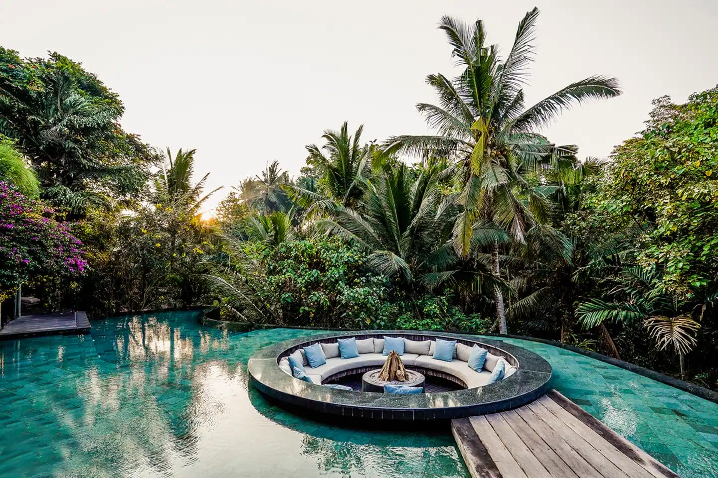 A circular sunken lounge with cushions and a fire pit is set in the middle of a pool, surrounded by tropical plants and palm trees.