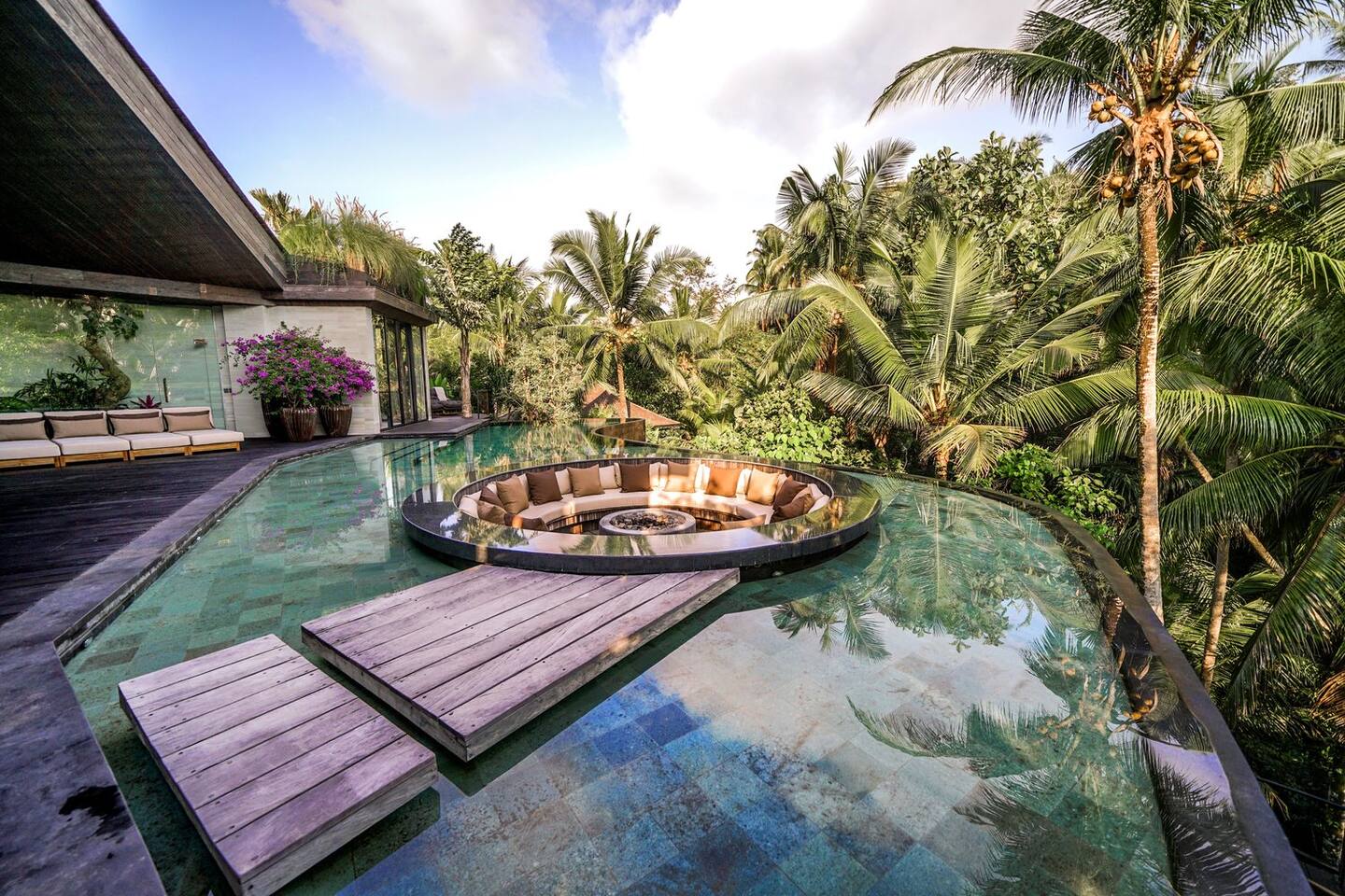 A circular sunken seating area with a fire pit is set in the middle of a reflective pool, surrounded by tropical trees and modern architecture.
