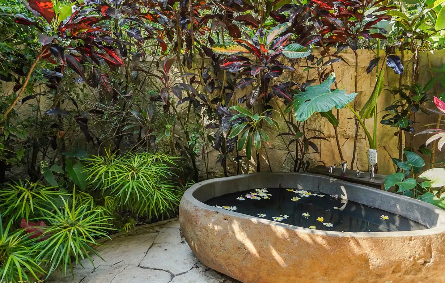 A stone bathtub filled with water and floating flowers, surrounded by lush tropical plants in an outdoor garden setting.