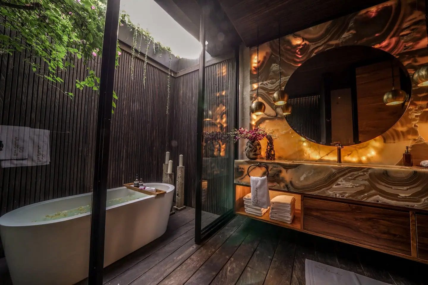Luxurious bathroom with a freestanding tub, large round mirror, modern sink, towels, wood flooring, and greenery outside a glass wall. Warm lighting creates a cozy ambiance.