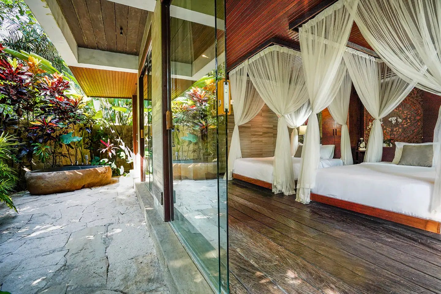 A bedroom with a canopy bed and sheer drapes is seen through a glass door, opening to a stone patio surrounded by lush tropical plants.
