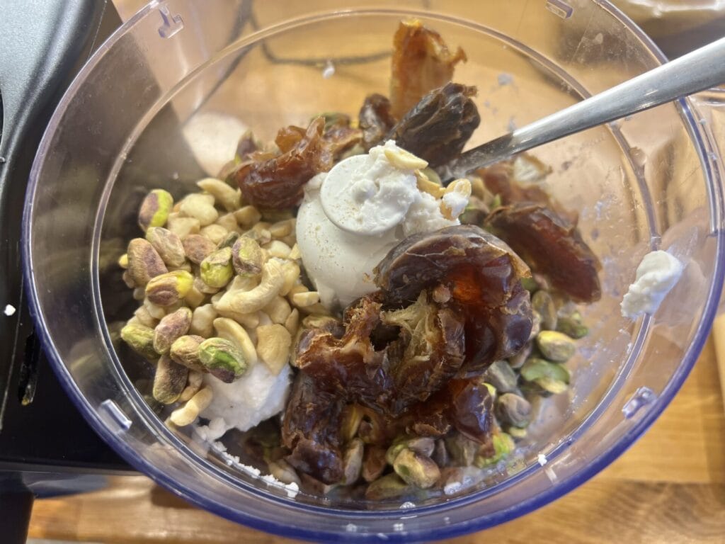 A food processor bowl containing pistachios, cashews, dates, and a dollop of white creamy ingredient, with a spoon inside.