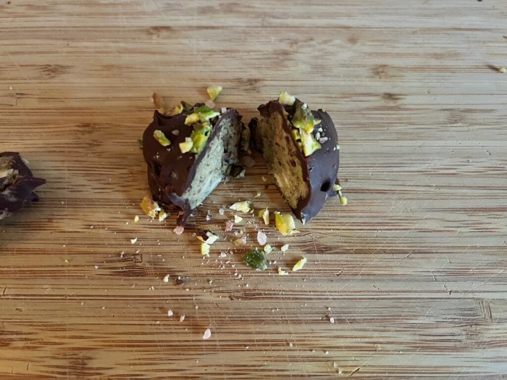 A chocolate-covered treat cut in half on a wooden surface, with chopped nuts sprinkled on top and crumbs scattered around.