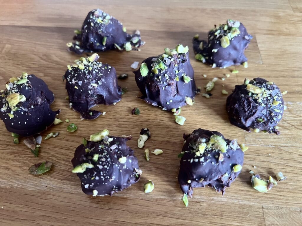 Eight Pistachio–Cashew Superfood Chocolate Bites, topped with chopped nuts, are arranged on a wooden surface.