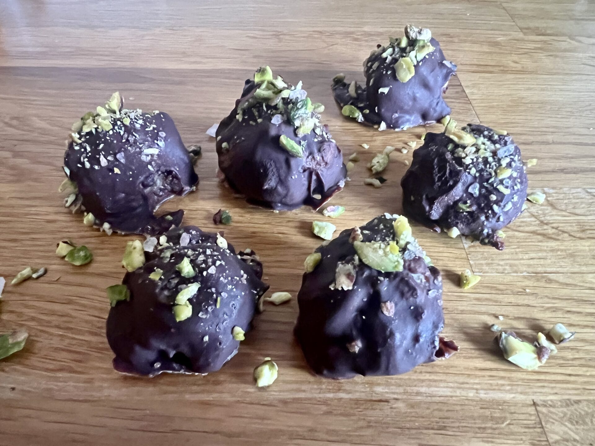 Six Pistachio–Cashew Superfood Chocolate Bites, coated in rich chocolate and topped with chopped nuts, are arranged on a wooden surface, with scattered nut pieces around them. Six Pistachio–Cashew Superfood Chocolate Bites, coated in rich chocolate and topped with chopped nuts, are arranged on a wooden surface, with scattered nut pieces around them.