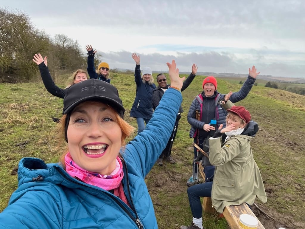 A group of people outdoors on a grassy hillside, smiling and raising their arms, with one person in front taking a selfie. Some are sitting on a wooden bench.