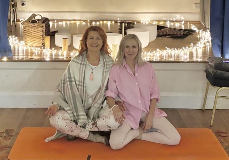 Two women sit cross-legged on an orange yoga mat in front of a raised platform decorated with string lights, bowls, and a basket.