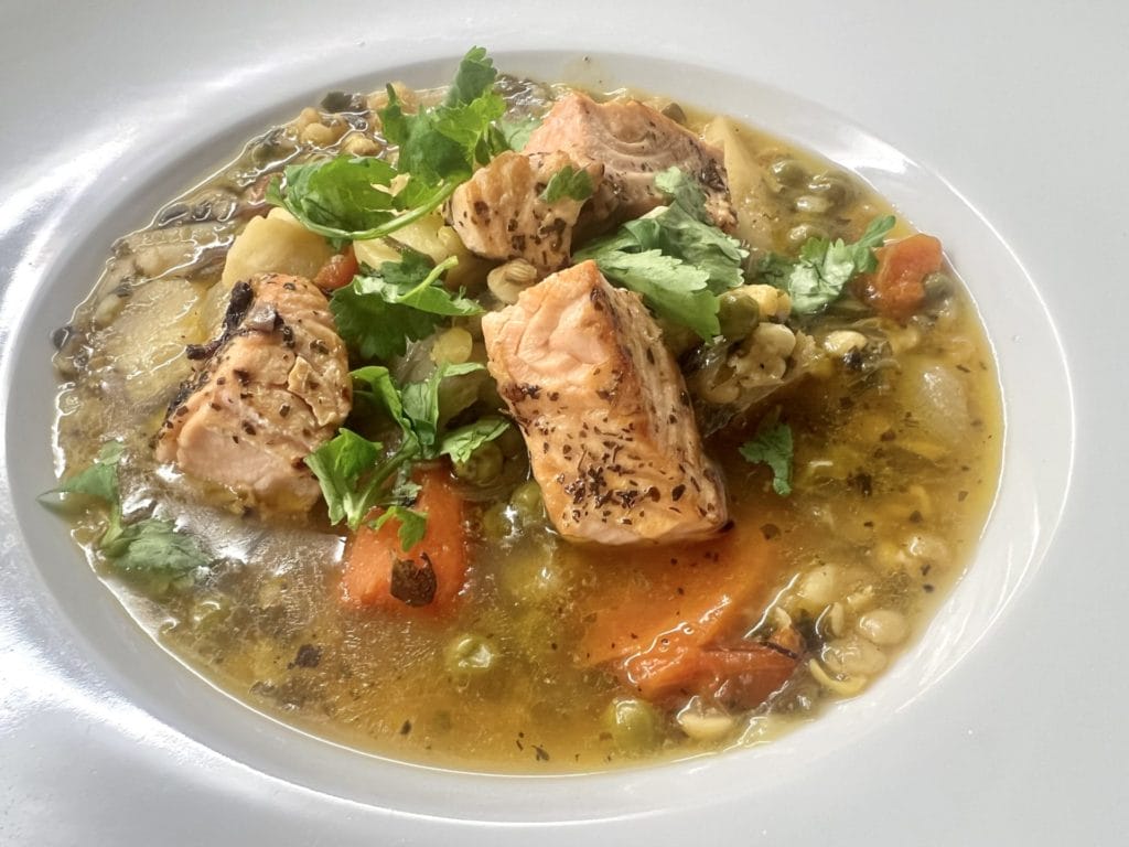 Chunks of cooked salmon, carrots, peas, and herbs in a broth, served in a white bowl.