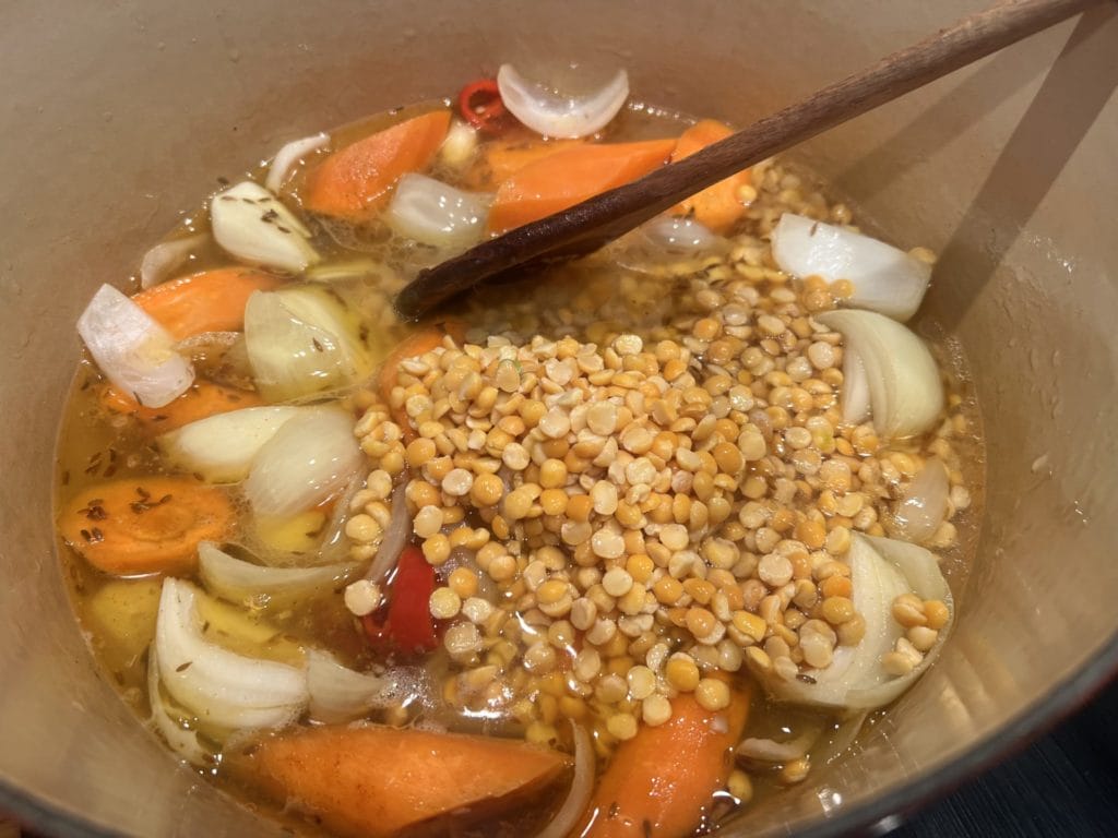 A pot containing yellow split peas, chopped carrots, onions, and spices being stirred with a wooden spoon while cooking in liquid for a Golden Lentil Stew with Baked Salmon – A Gut & Brain Warming Delight.