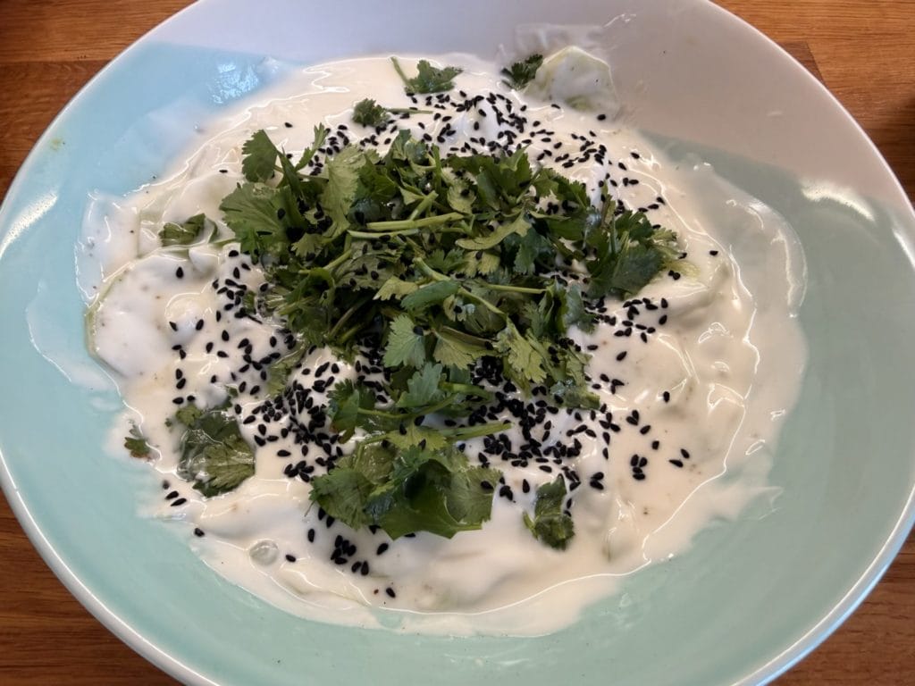A bowl of yoghurt garnished with fresh coriander leaves and black sesame seeds, on a wooden surface.