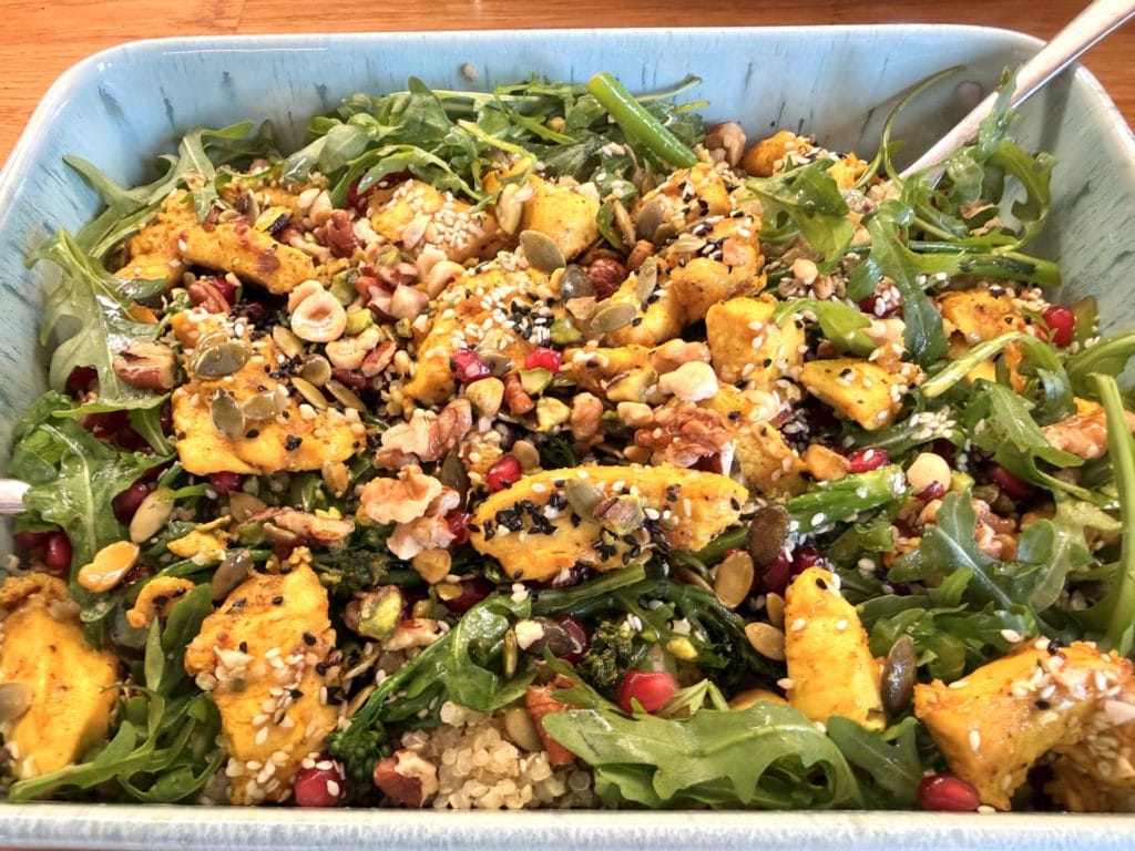 A salad with rocket, quinoa, grilled chicken pieces, pomegranate seeds, mixed nuts, and seeds in a blue dish.