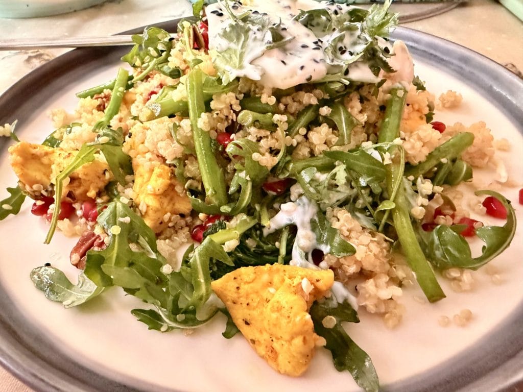 A plate of quinoa salad with rocket, green beans, pomegranate seeds, tofu pieces, and a dollop of yoghurt topped with herbs and black sesame seeds.