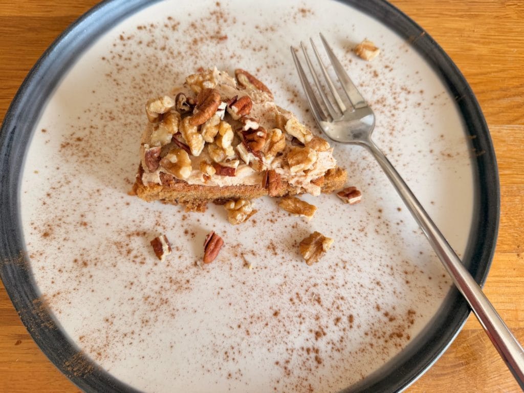 A slice of cake topped with chopped nuts on a plate dusted with cinnamon, with a fork placed beside the cake.